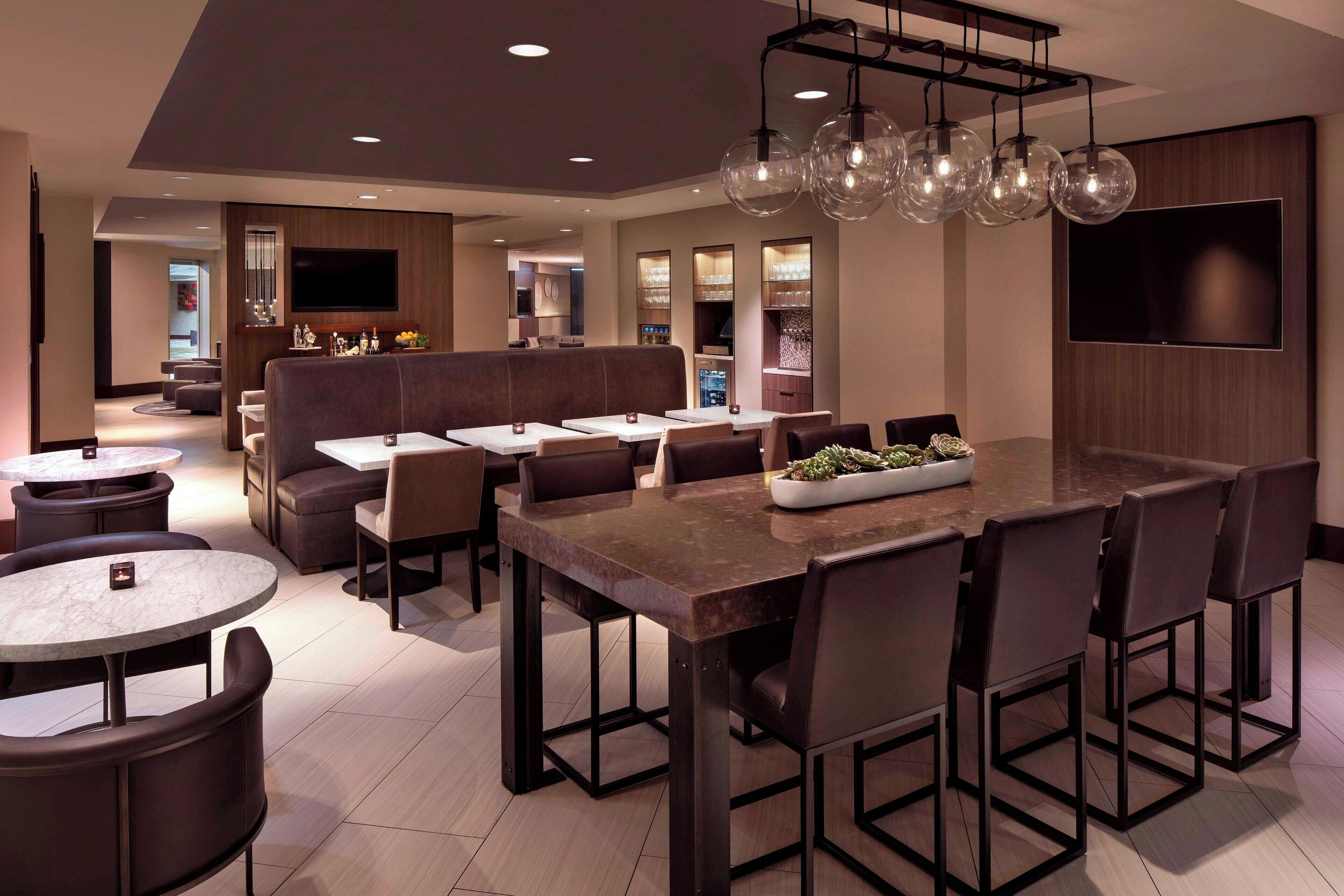 A modern restaurant with a central bar area, multiple seating options including round tables and booths, and a large TV screen on the wall.