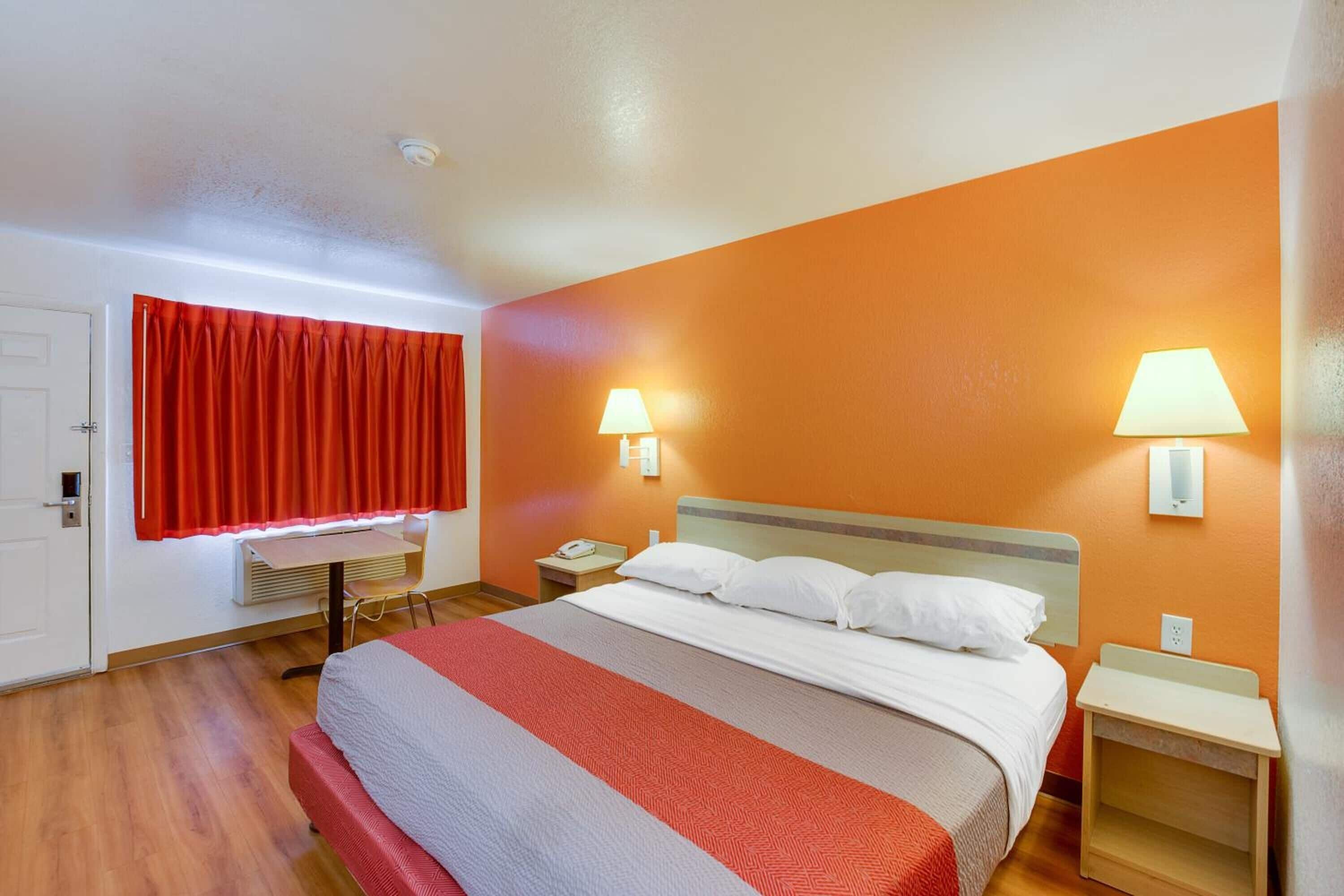 A hotel room with a large bed, two bedside tables, a window with red curtains, and two wall-mounted lamps.