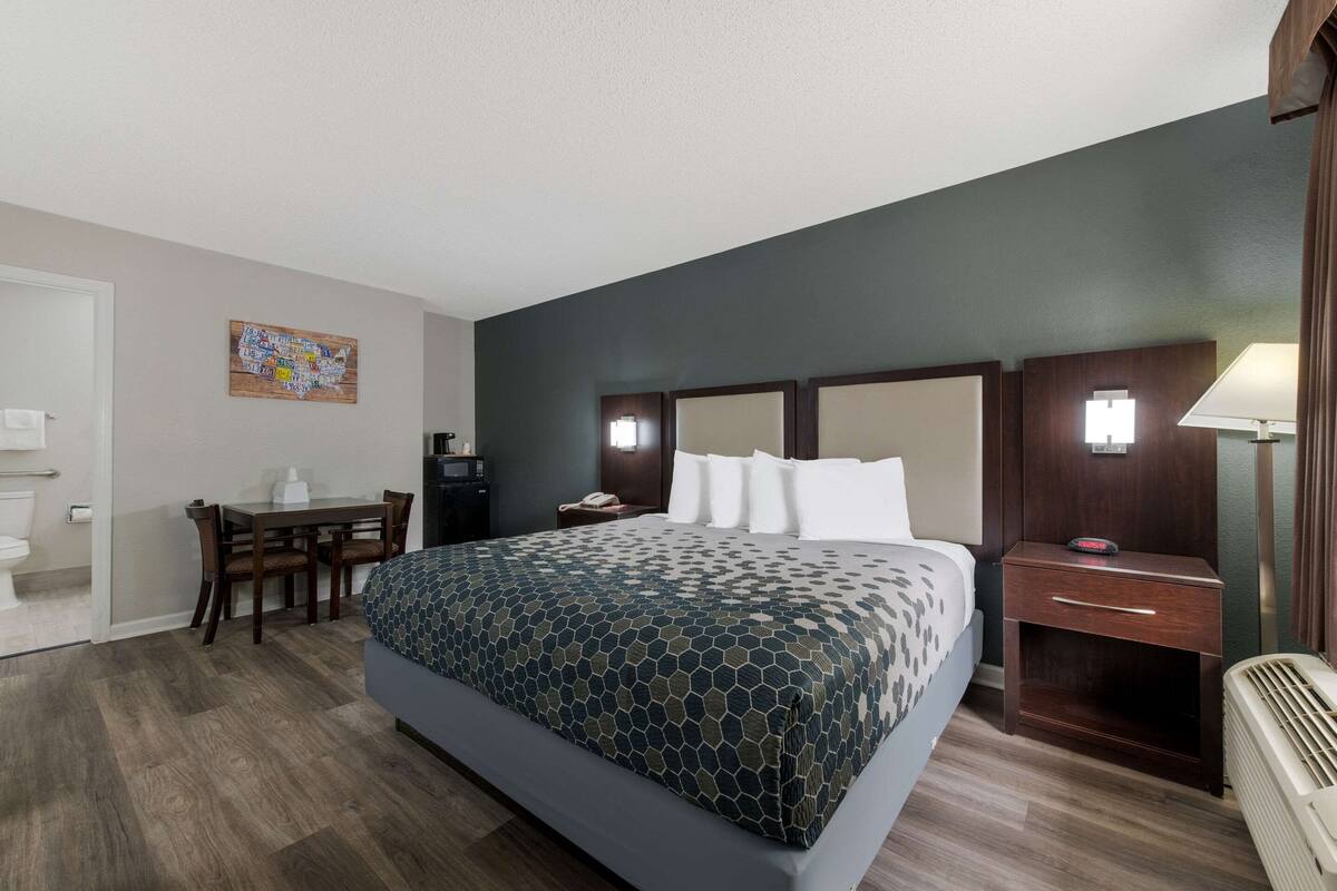 Ein Hotelzimmer mit einem großen Bett, einem Schreibtisch, einem Stuhl, einem Nachttisch, einer Lampe und einem an der Wand montierten Fernseher.