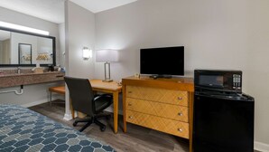 Habitación de hotel con cama, escritorio y silla, televisor, microondas, cómoda y baño con lavamanos y espejo.