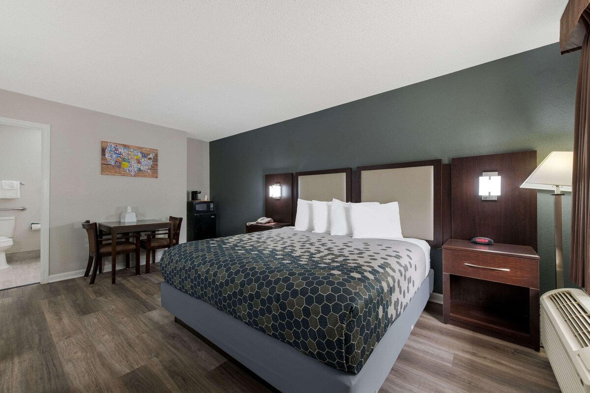 A hotel room with a large bed, a desk, and a chair. There is a nightstand with a lamp and a phone.