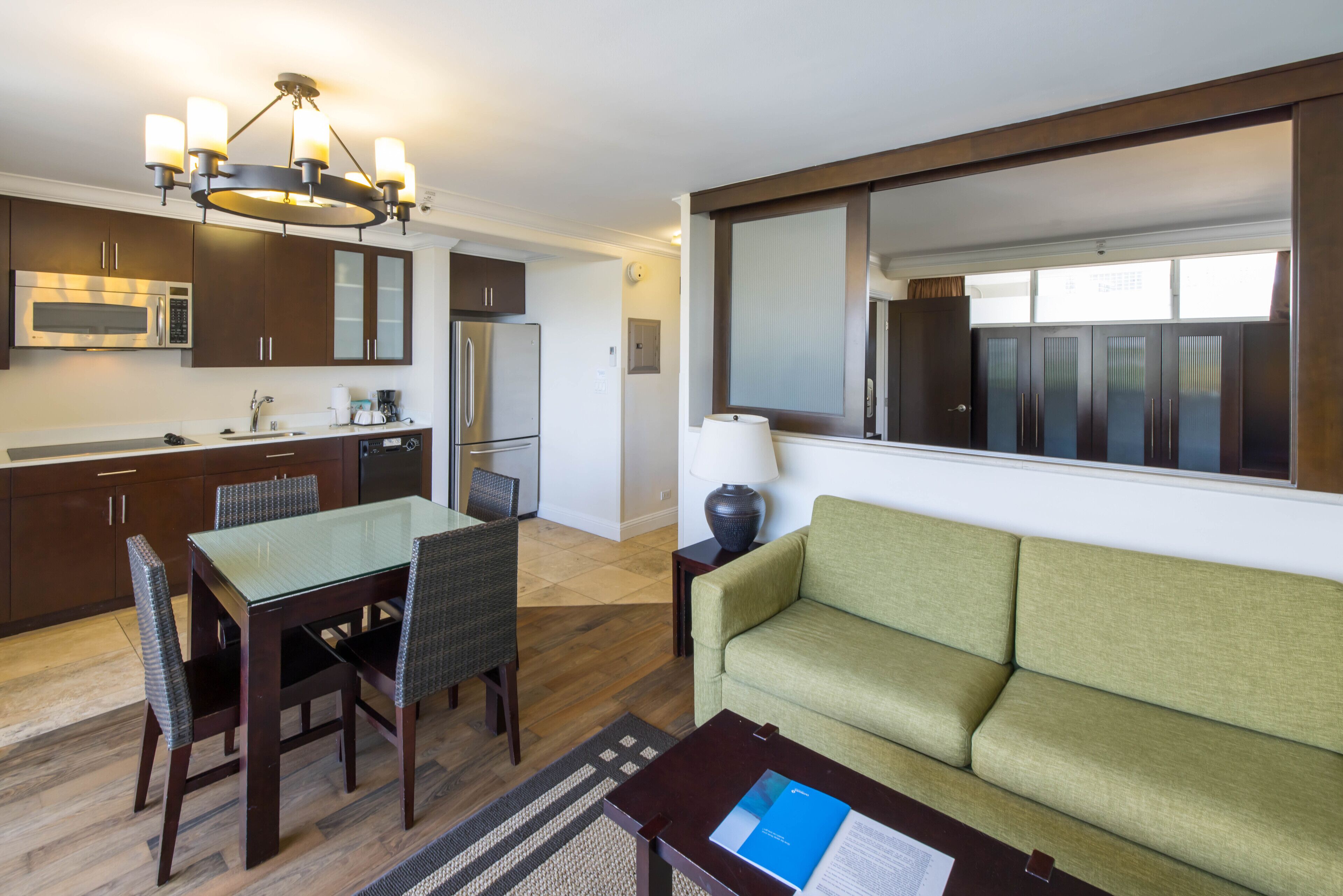 A modern kitchen with dark wood cabinets, a stainless steel microwave, and a built-in oven. There is a dining area with a glass table and wicker chairs. A green sofa is placed in a corner with a lamp on a side table.