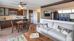 A modern kitchen with dark cabinets, a microwave, and a refrigerator. There is a dining area with a wooden table and chairs. A white sofa with patterned cushions is in the living area. - Regency on Beachwalk Waikiki by OUTRIGGER (Honolulu)