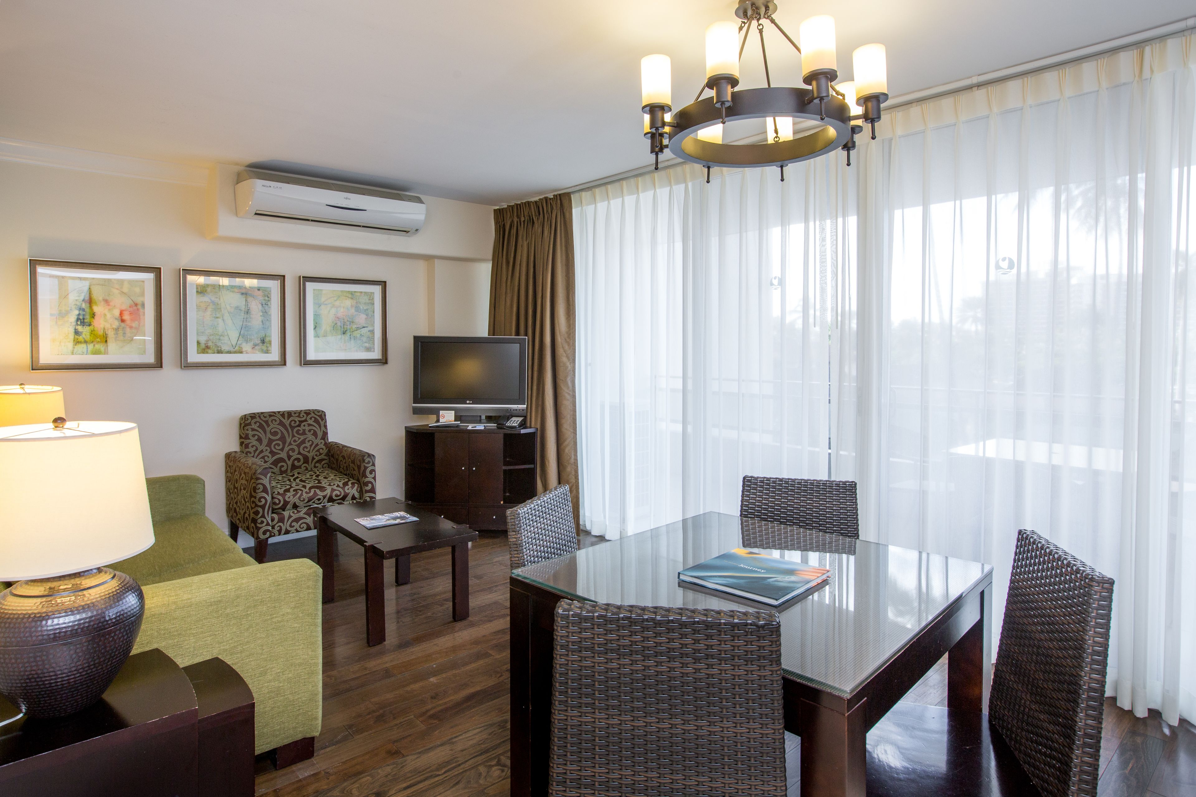 A hotel room with a glass dining table, wicker chairs, a flat-screen TV, a sofa, and a lamp.