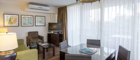 A hotel room with a glass dining table, wicker chairs, a flat-screen TV, a sofa, and a lamp.