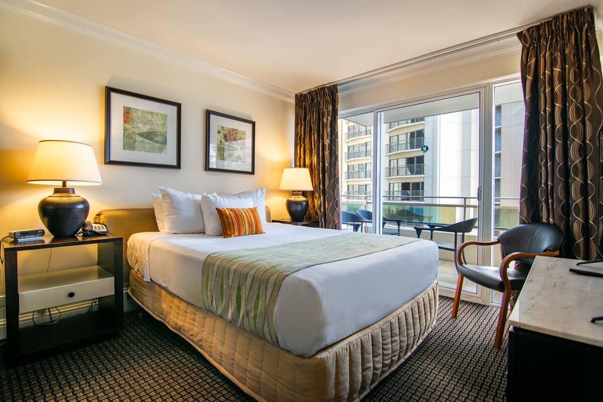 A hotel room with a bed, two bedside lamps, a nightstand, a chair, a table, and a view of a building through a window.