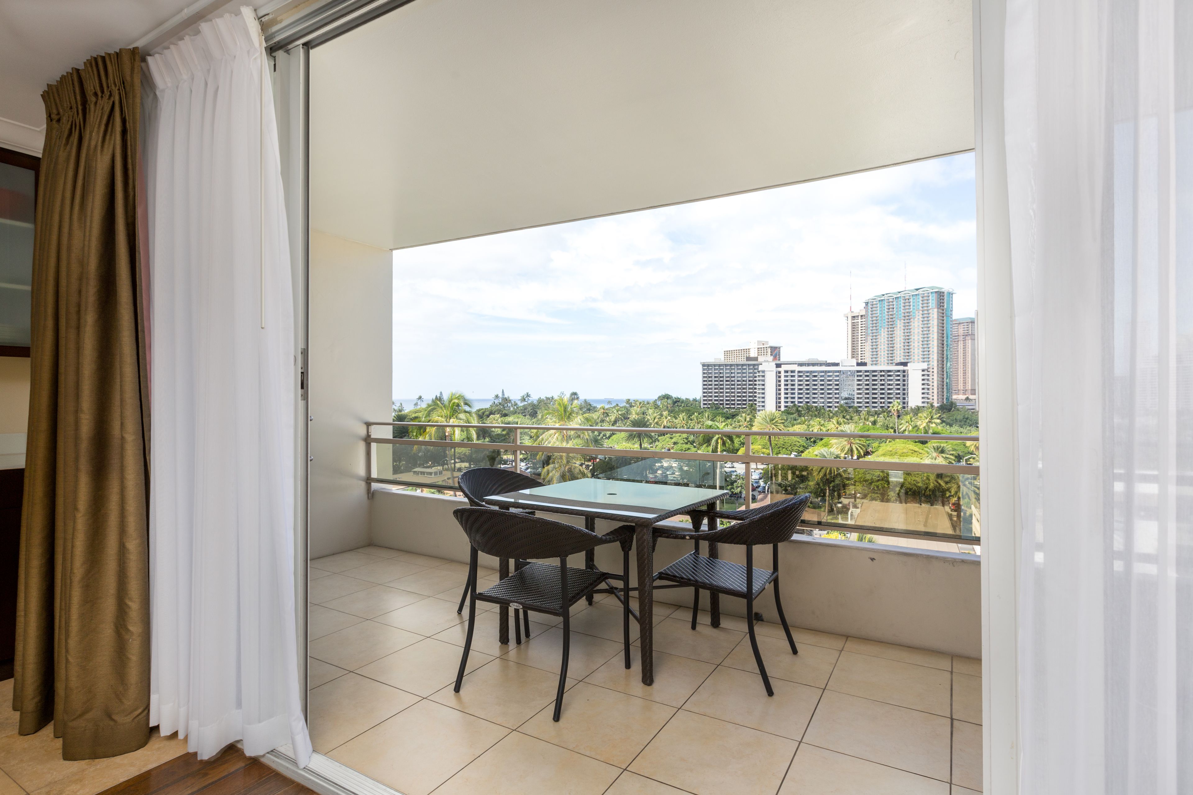 A balcony with a glass table and chairs, a view of city buildings, and a green landscape.