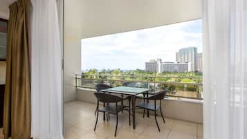 A balcony with a glass table and chairs, a view of city buildings, and a green landscape.