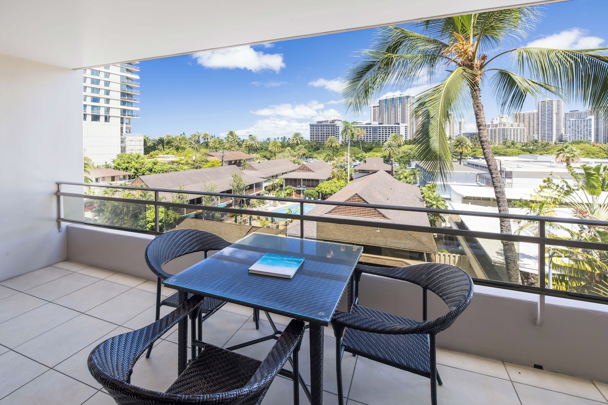 A balcony with a table and chairs, overlooking a cityscape with buildings and palm trees.