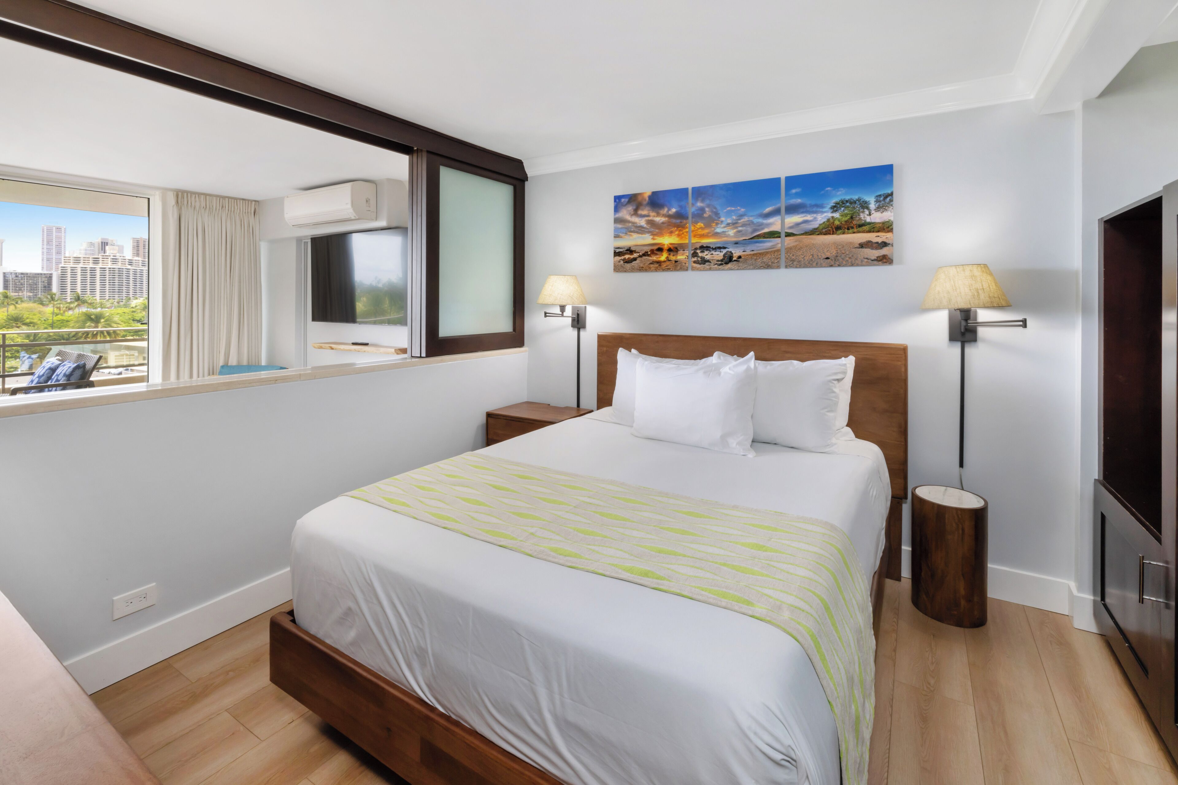 Una habitación de hotel con una cama grande, un ventanal con vistas a edificios, dos lámparas de noche y un cuadro en la pared.