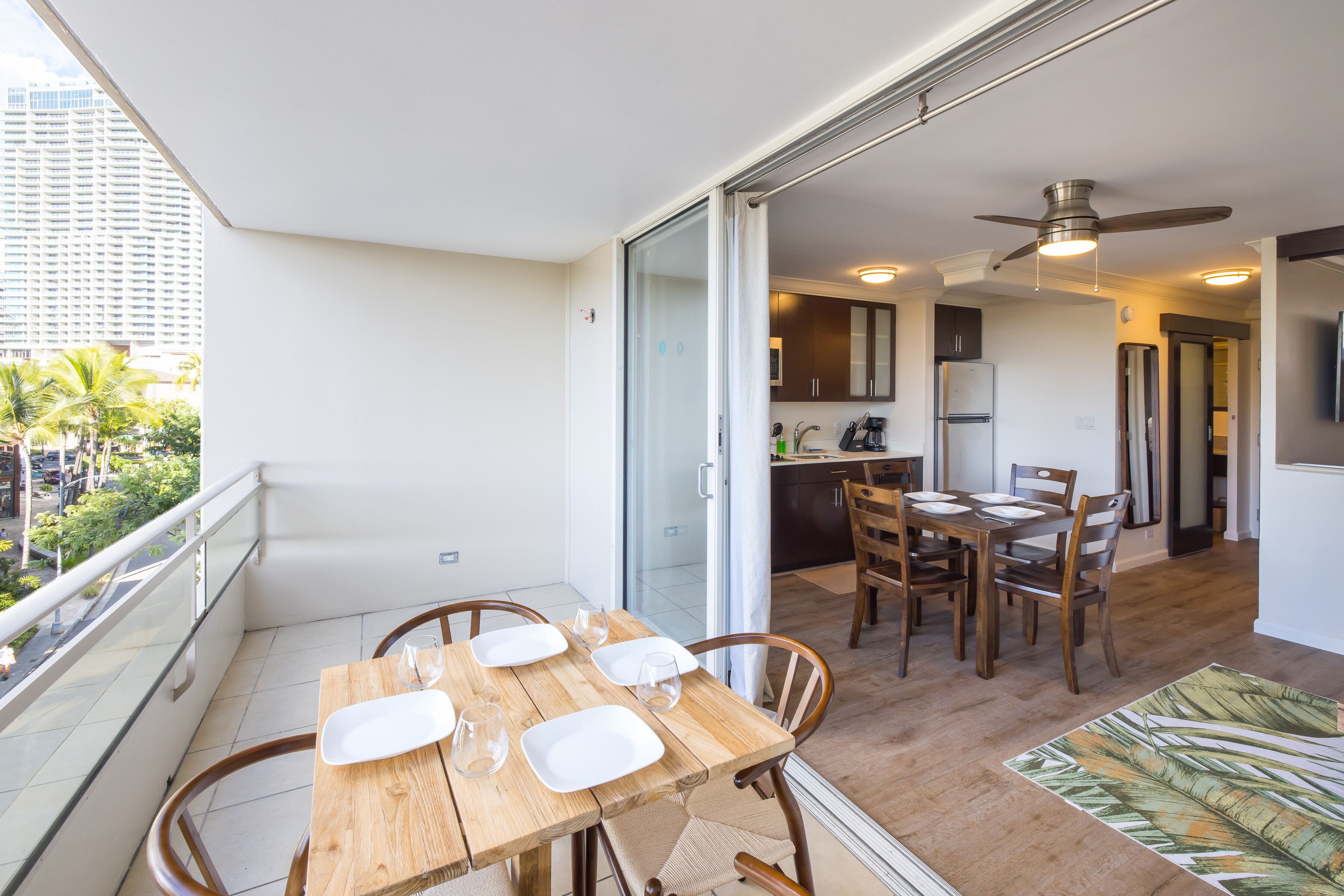 Un balcón con mesa y sillas para cuatro, una cocina al fondo y vistas a un edificio alto y palmeras.