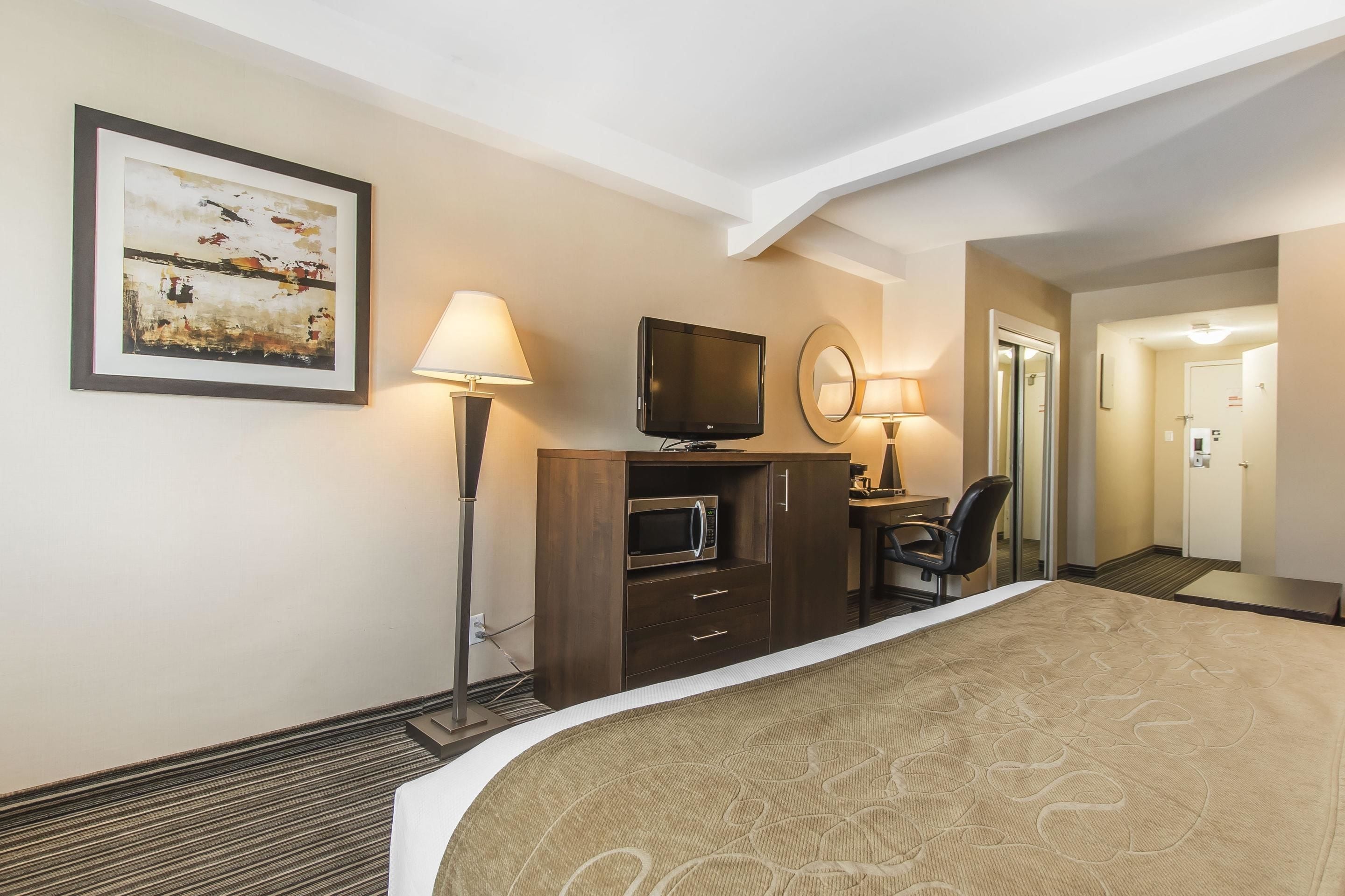 A hotel room with a bed, a desk with a computer, a television, a microwave, a lamp, and a framed picture on the wall.