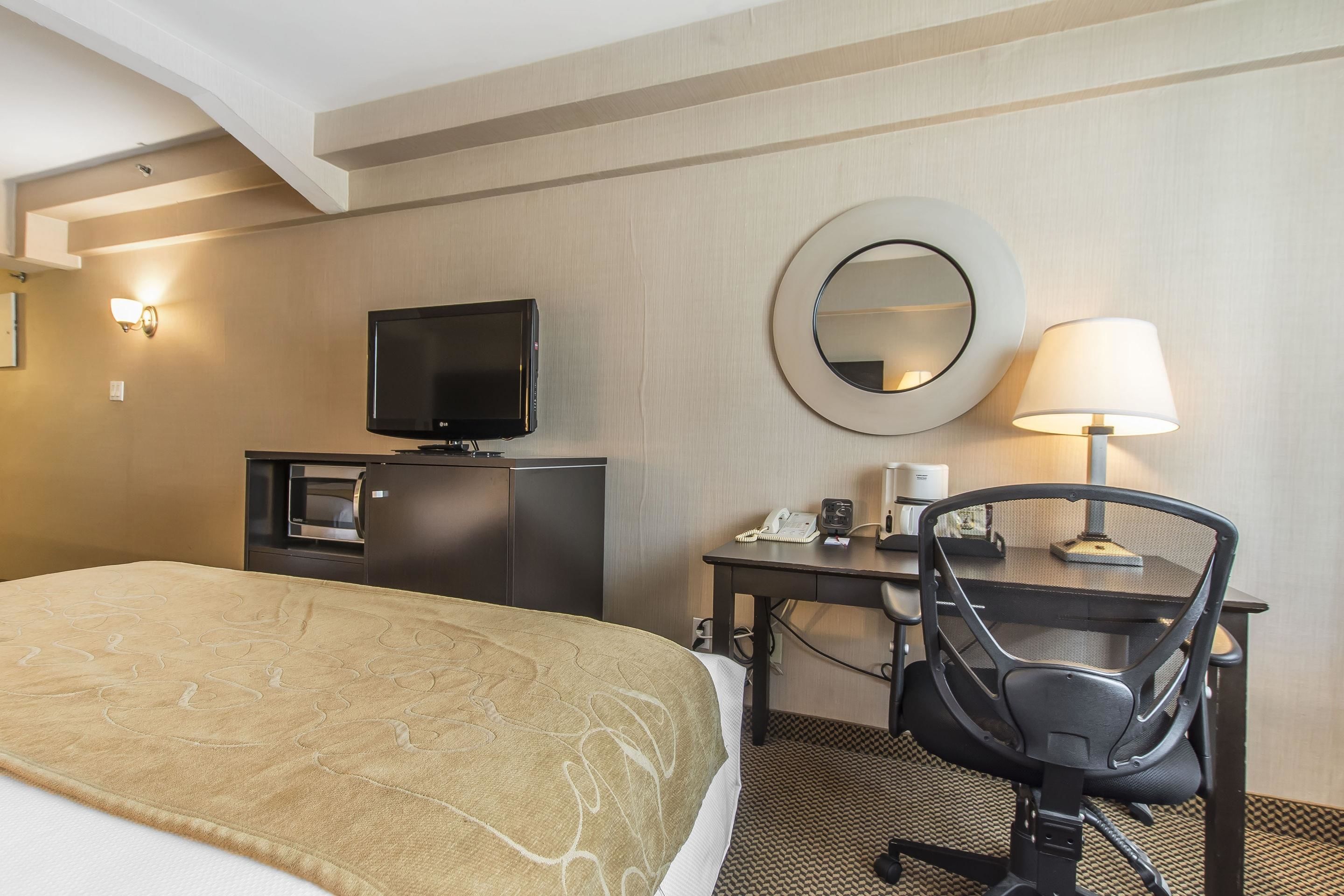 A hotel room with a bed, a desk with a chair, a television, a mirror, and a lamp.