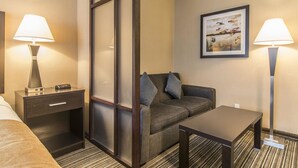 Una habitación de hotel con cama, mesita de noche con lámpara, sofá, una pequeña mesa y un cuadro en la pared.