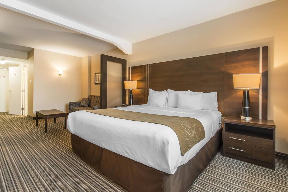 Una habitación de hotel con una cama grande, una mesita de noche, dos lámparas, una silla y una pequeña mesa.