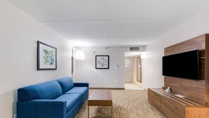A modern hotel room with a blue sofa, a wooden TV stand, and a small table. - Holiday Inn Express Naperville by IHG (Naperville)