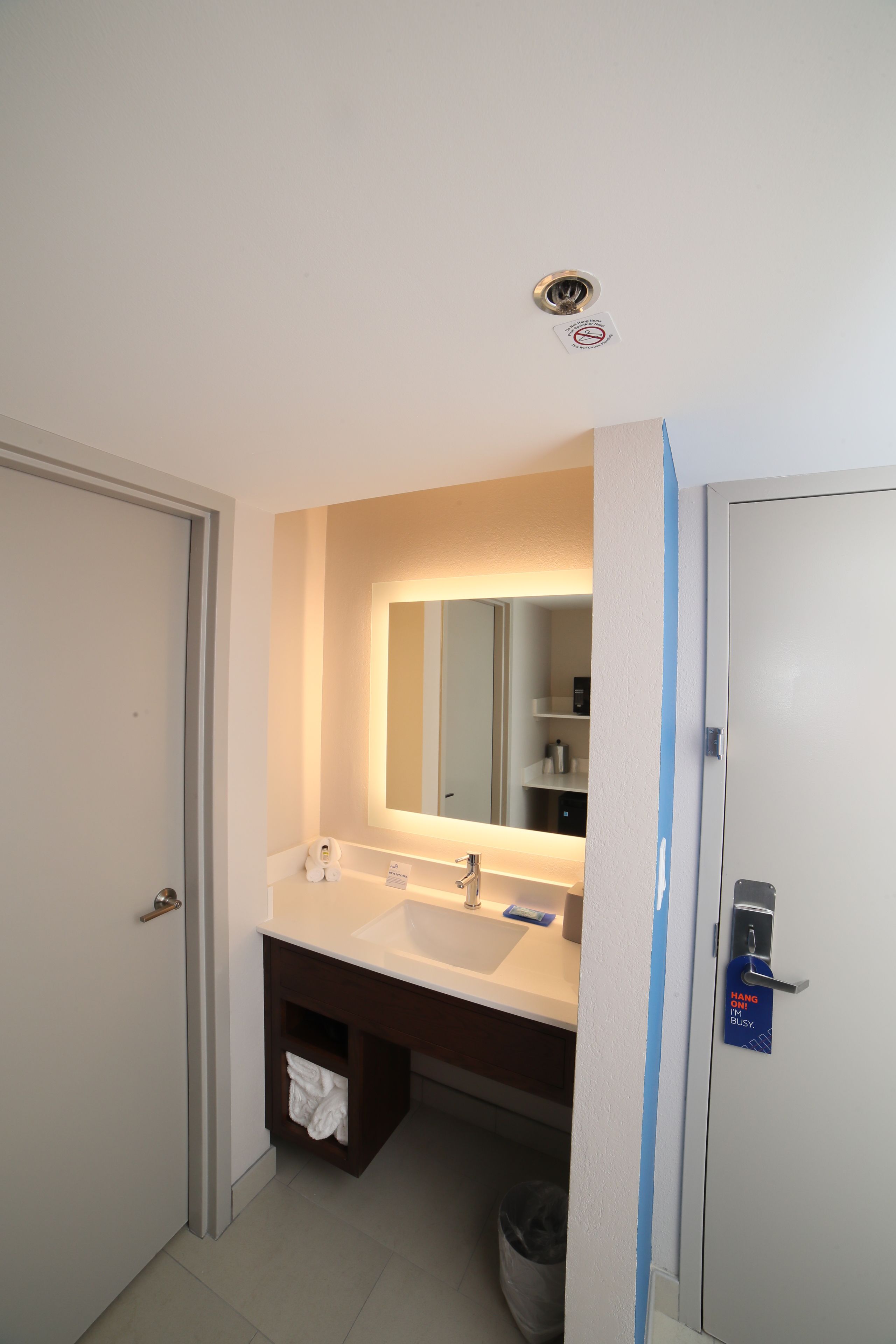 A hotel bathroom with a white sink, mirror, and a blue sign on the door.