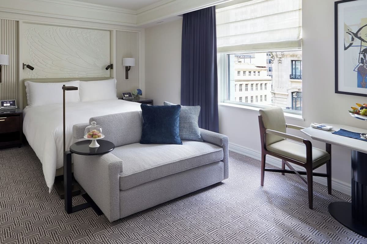 A hotel room with a bed, a sofa, a desk, and a chair. There is a window with a view of buildings.