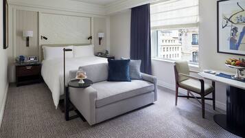Habitación de hotel con cama, sofá, escritorio y silla. Se ve una ventana con vistas a edificios.