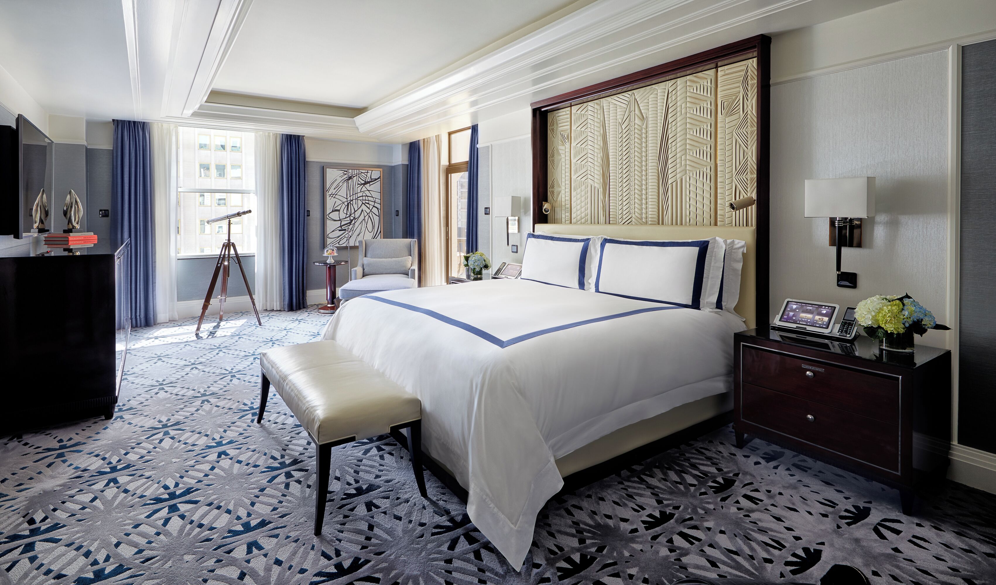 A hotel room with a large bed, a desk with a computer, a chair, a desk lamp, and a window with curtains.