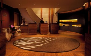 A reception area with a wooden floor, a large rug, wooden reception desks, and a wall with a display shelf.
