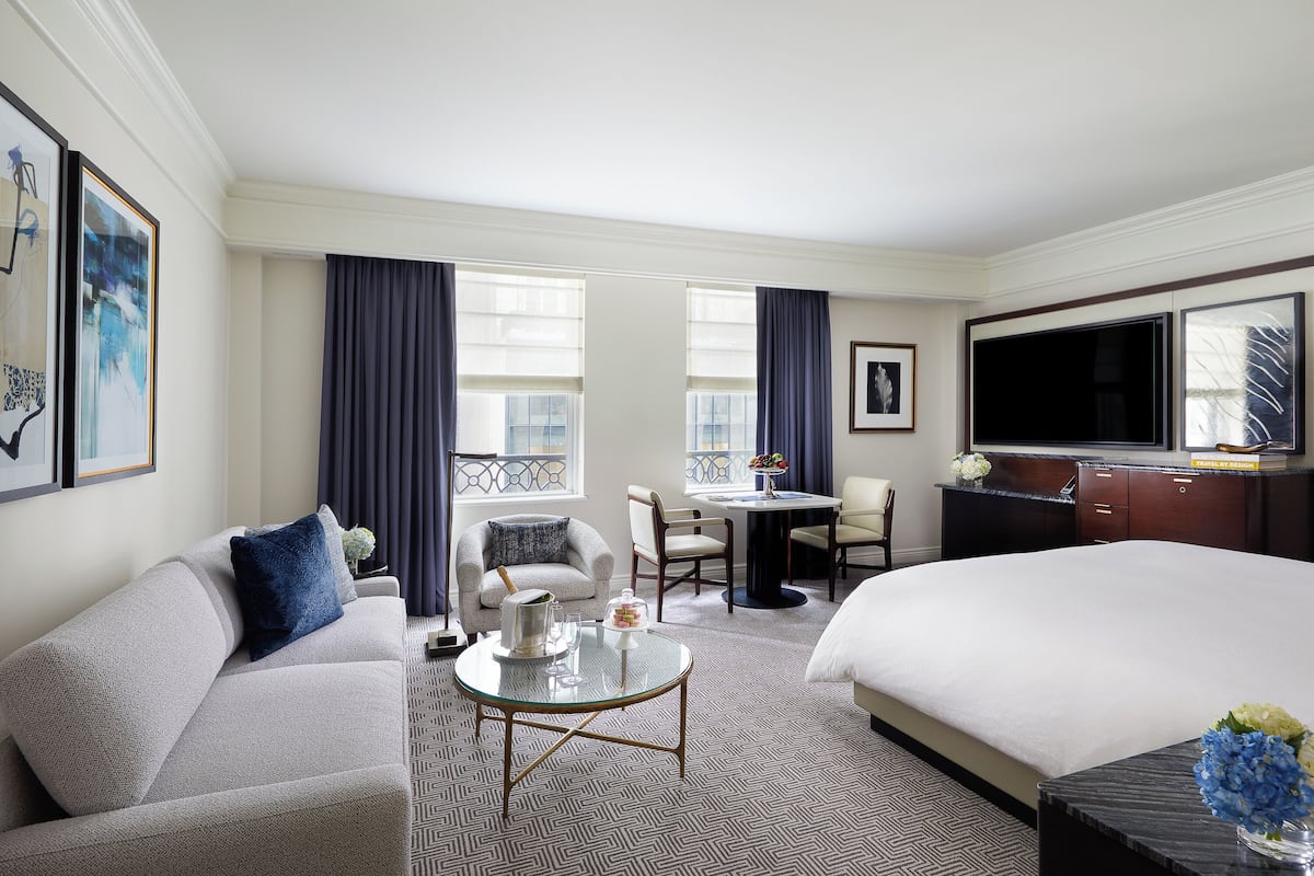 A hotel room with a large bed, a sofa, a dining table with chairs, a TV, and artwork on the walls.