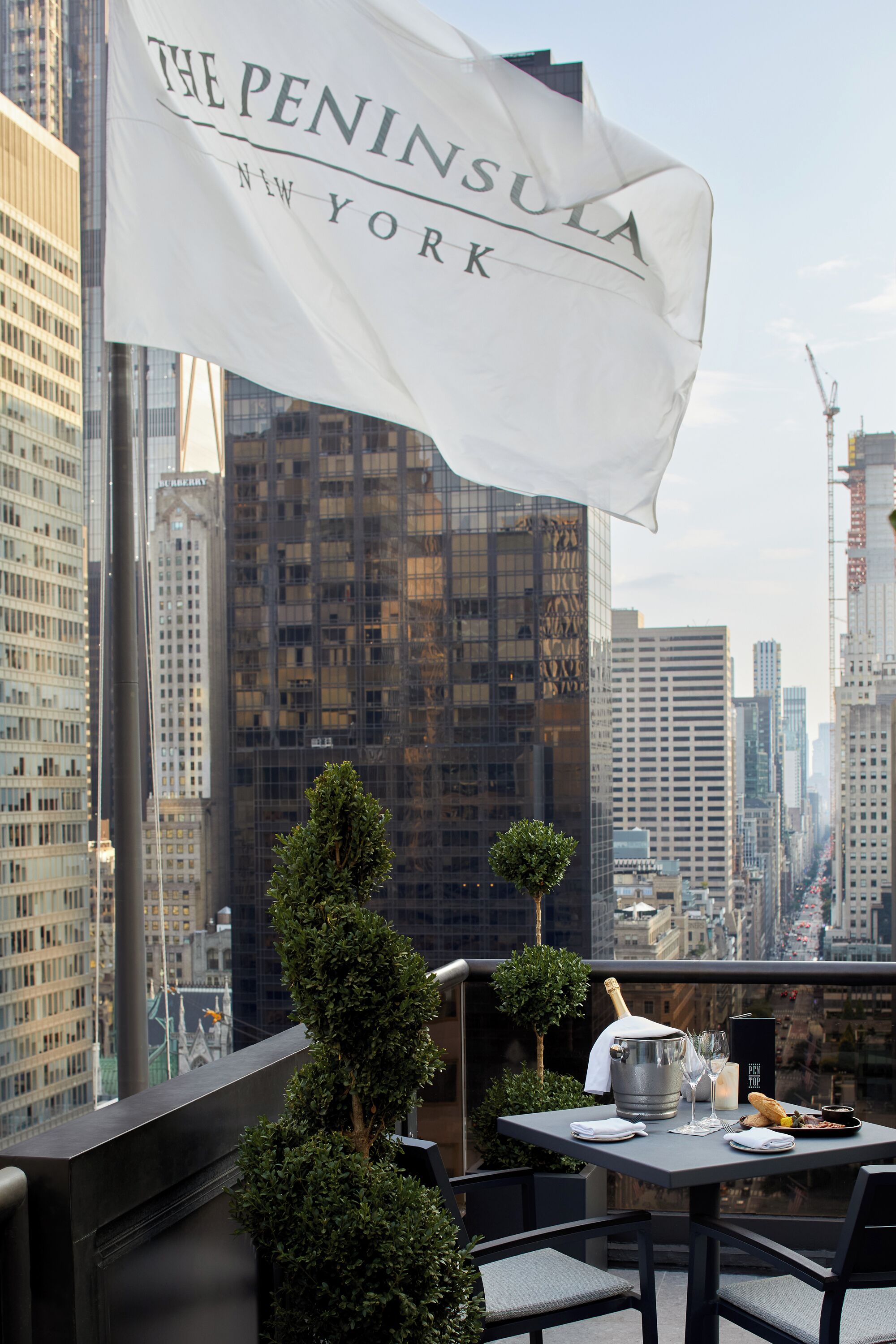 A rooftop terrace with a table set for dining, a champagne bottle, and a flag reading "The Peninsula New York".