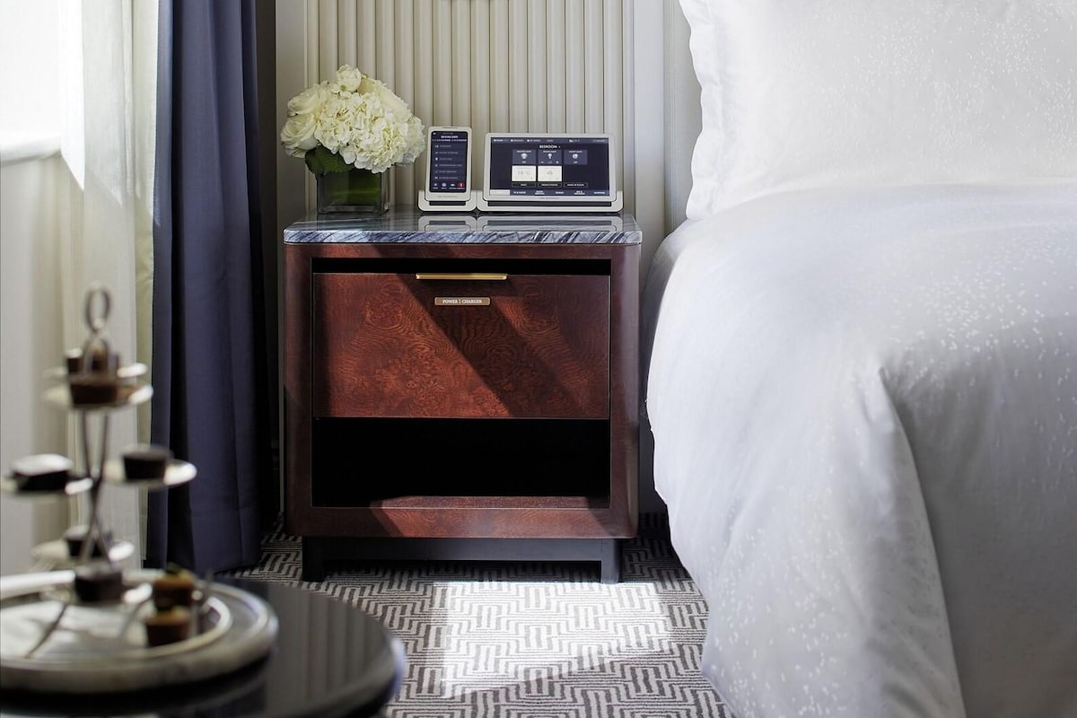 Habitación de hotel con cama, mesita de noche con lámpara, regleta y bandeja decorativa.