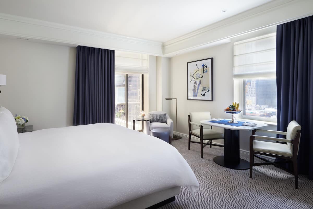 A hotel room with a large bed, a desk with a chair, a small table, and a view of the city through large windows.