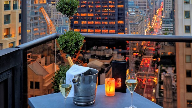 Ein Tisch mit einer Champagnerflasche, zwei Gläsern und einer Speisekarte auf einem Balkon mit Blick auf eine Stadtlandschaft, hohe Gebäude und eine Flagge.