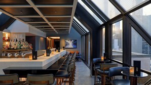 A modern bar area with a long counter, bar stools, and a view of the city through large windows. - The Peninsula New York (New York)