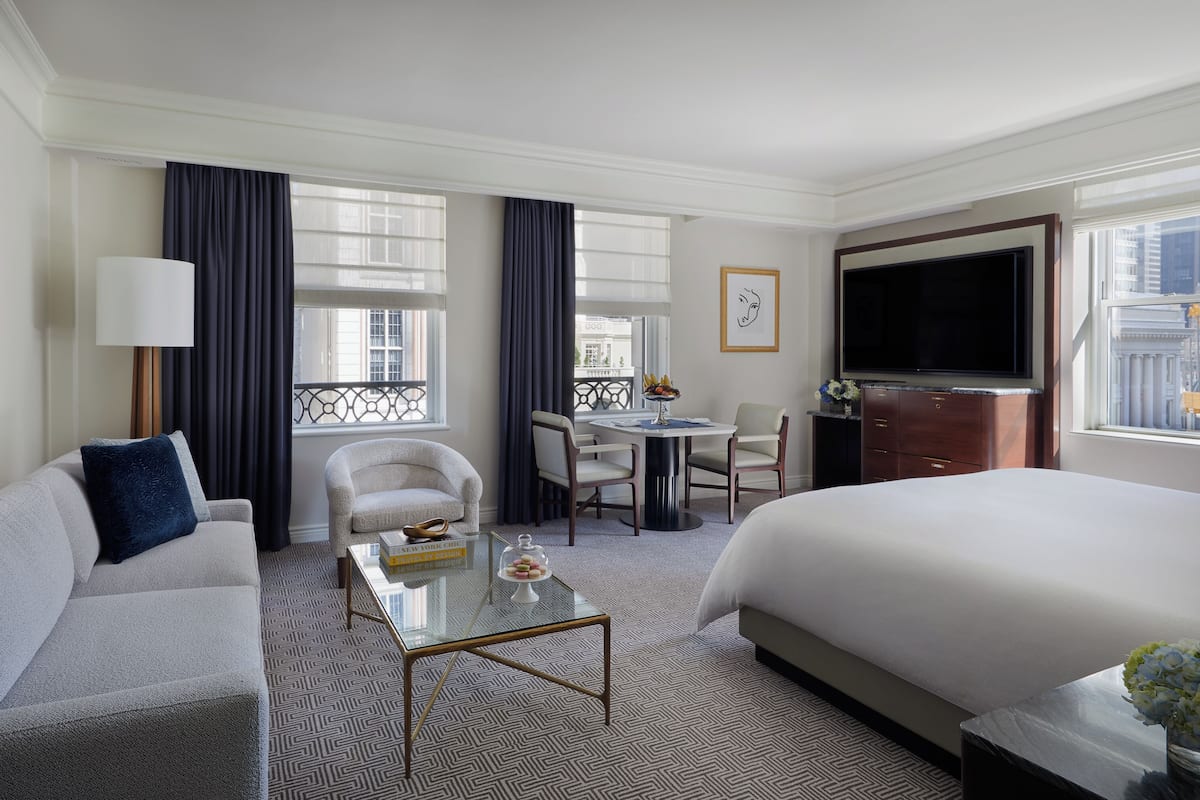 A hotel room with a bed, a sofa, a dining table with chairs, a TV, and artwork on the walls.