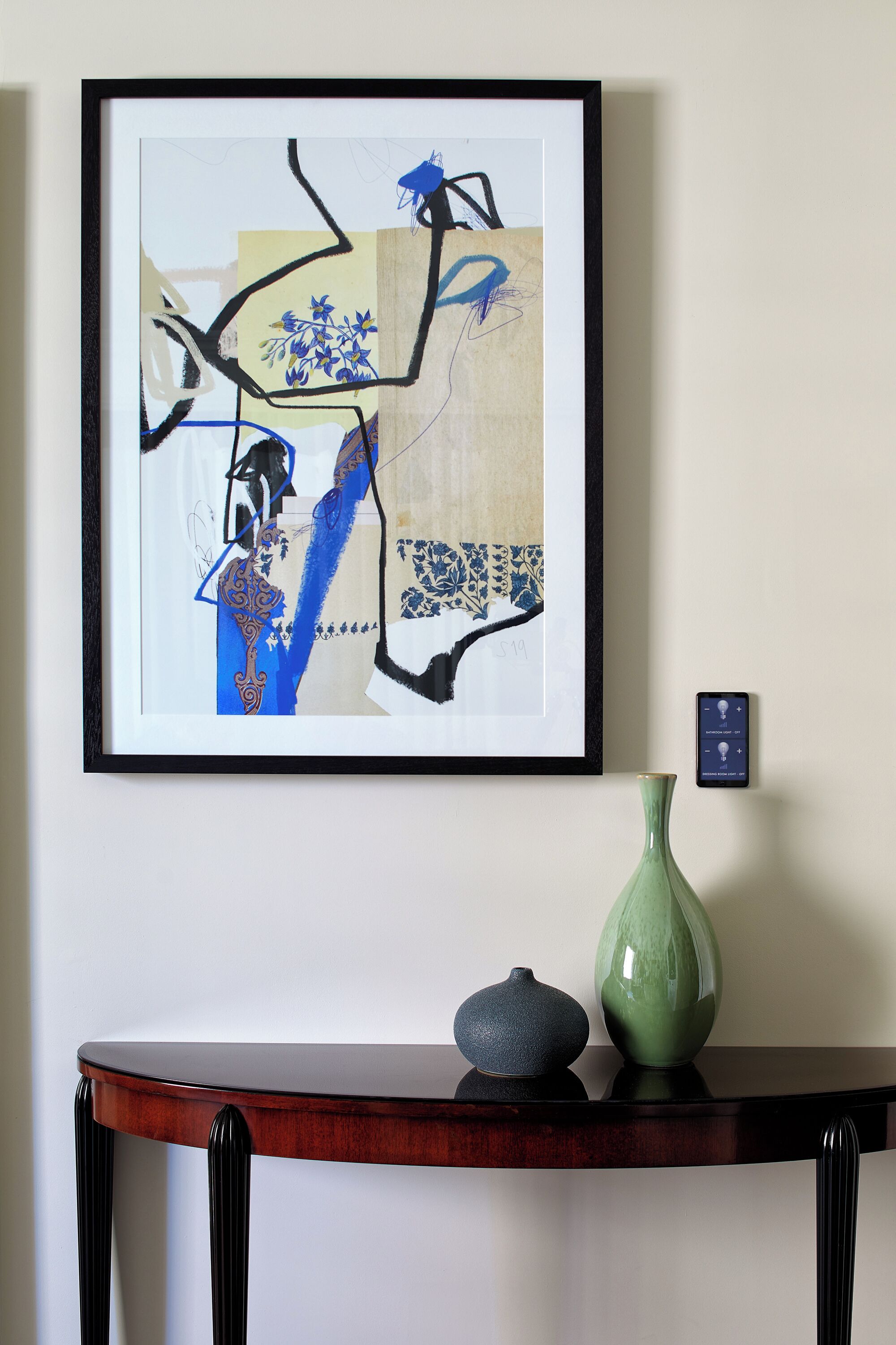 A framed abstract painting on a wall, a small table with two vases, and a wall-mounted light switch.