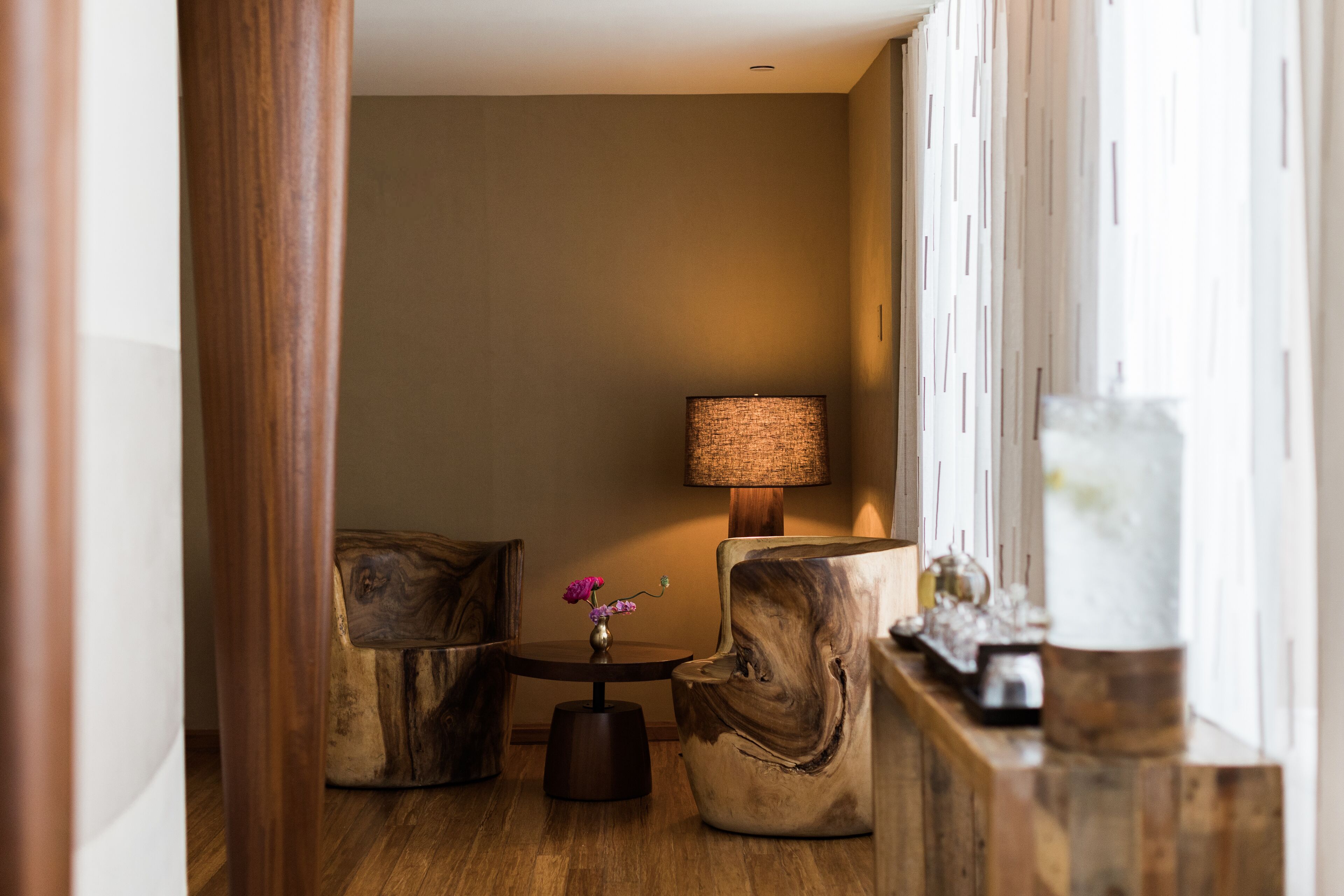 Um quarto com chão de madeira, uma mesa redonda com um vaso de flores, duas cadeiras de madeira, um abajur e um objeto decorativo sobre um aparador.
