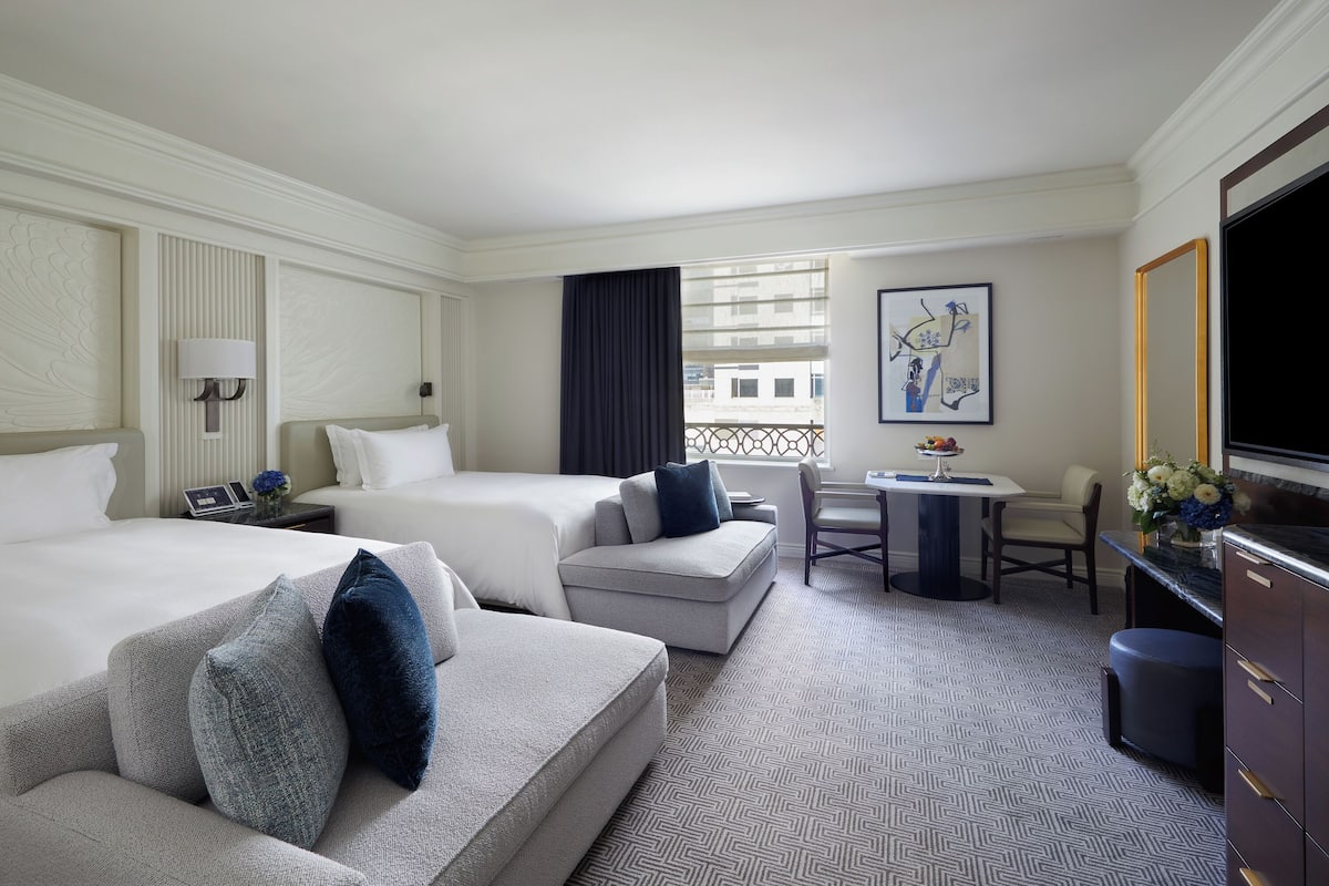 A hotel room with two beds, a desk, a chair, a TV, and a window with a view of buildings.