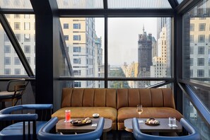 A modern interior with a large window offering a city view, a brown leather sofa, and two small tables with drinks and snacks.