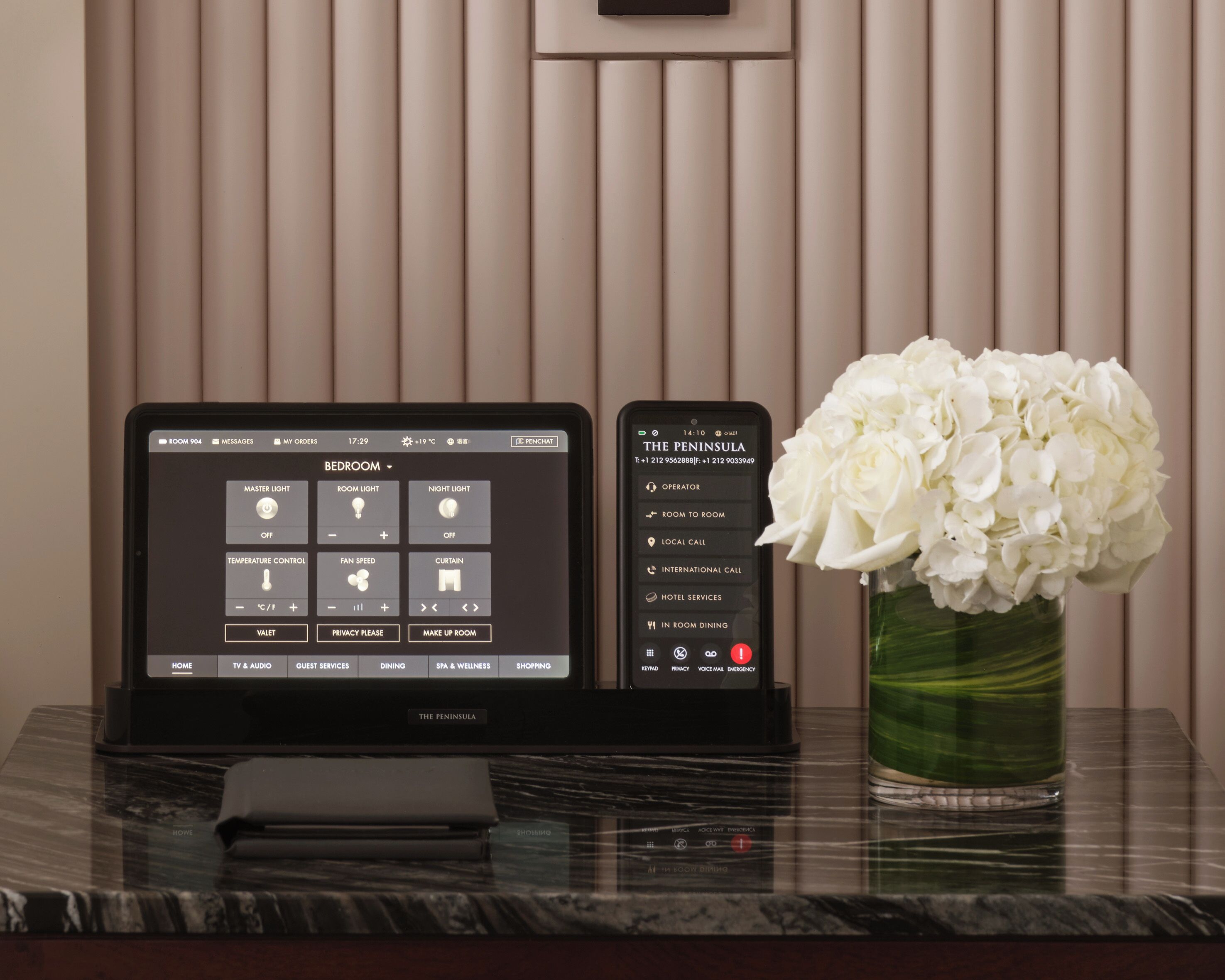 A tablet displaying room control options, a smartphone with hotel contact information, and a vase of white flowers on a marble table.