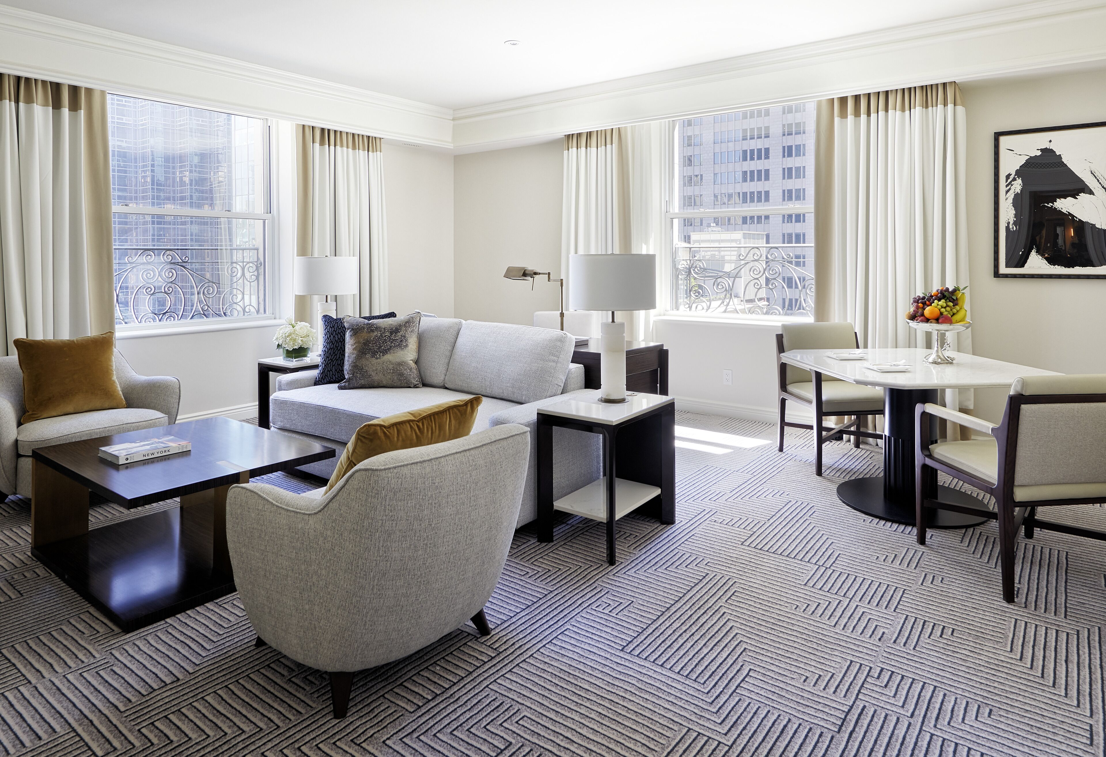 A modern hotel room with a sofa, armchair, dining table, and a view of the city through large windows.