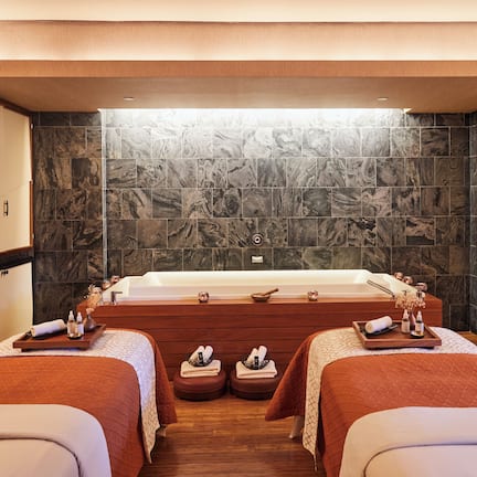 A spa room with a large bathtub, two massage tables, and a stone wall.