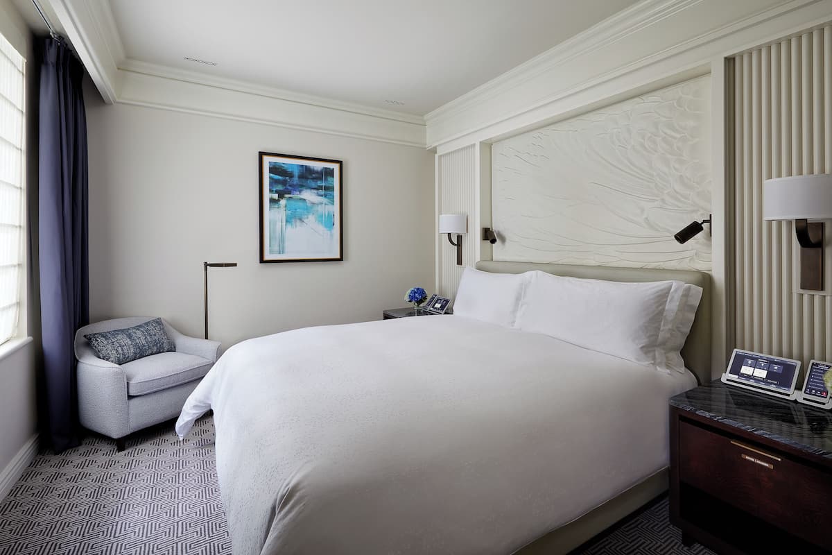A hotel room with a large bed, a bedside table with a lamp, a chair, and a painting on the wall.