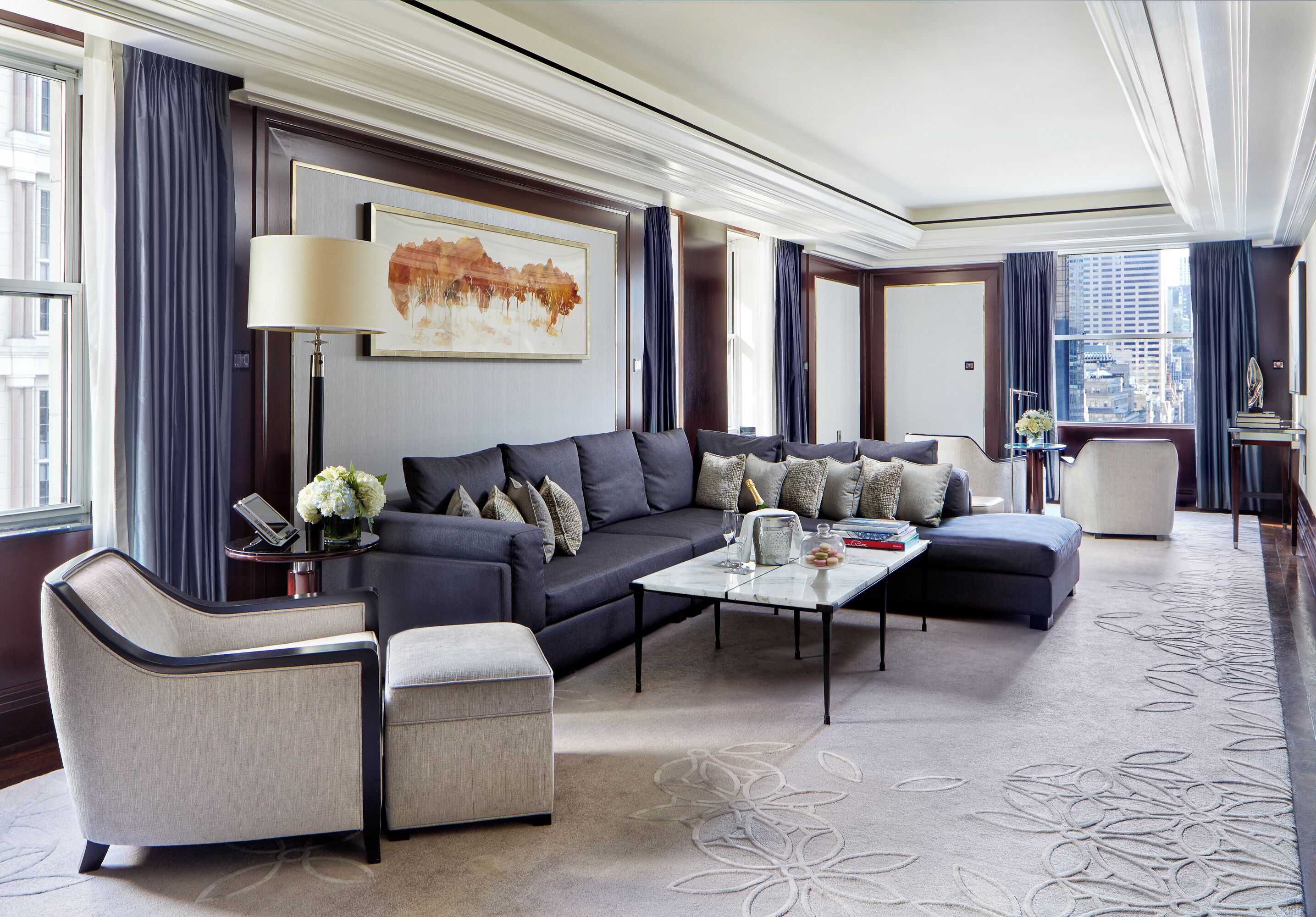 Ein modernes Hotelzimmer mit einer dunkelgrauen Sofagarnitur, einem passenden Sessel, einem Couchtisch und einem großen Fenster mit Blick auf die Stadt.