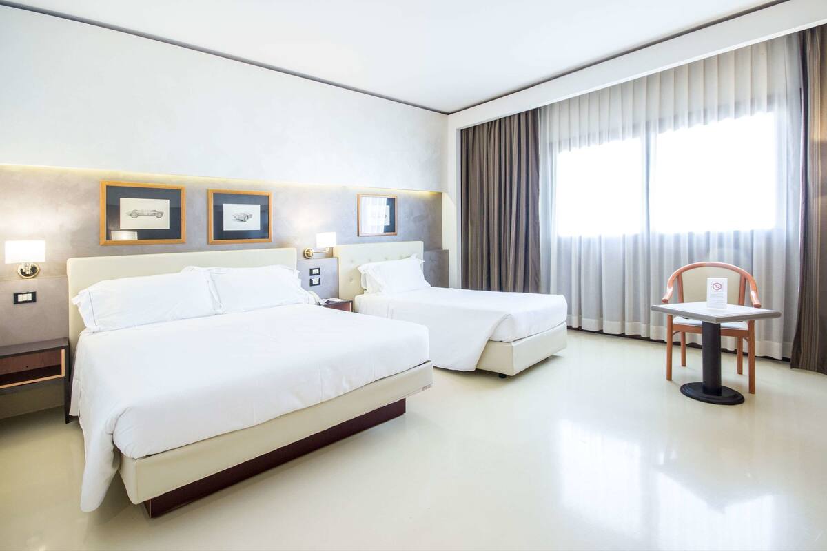Ein Hotelzimmer mit zwei Betten, einem Tisch und einem Stuhl. An der Wand befinden sich Bilder in Rahmen und ein Fenster mit Vorhängen.