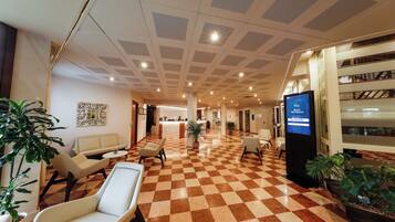 Eine Hotellobby mit Schachbrettmuster-Boden, Sitzbereichen, einer Rezeption und einem digitalen Bildschirm.