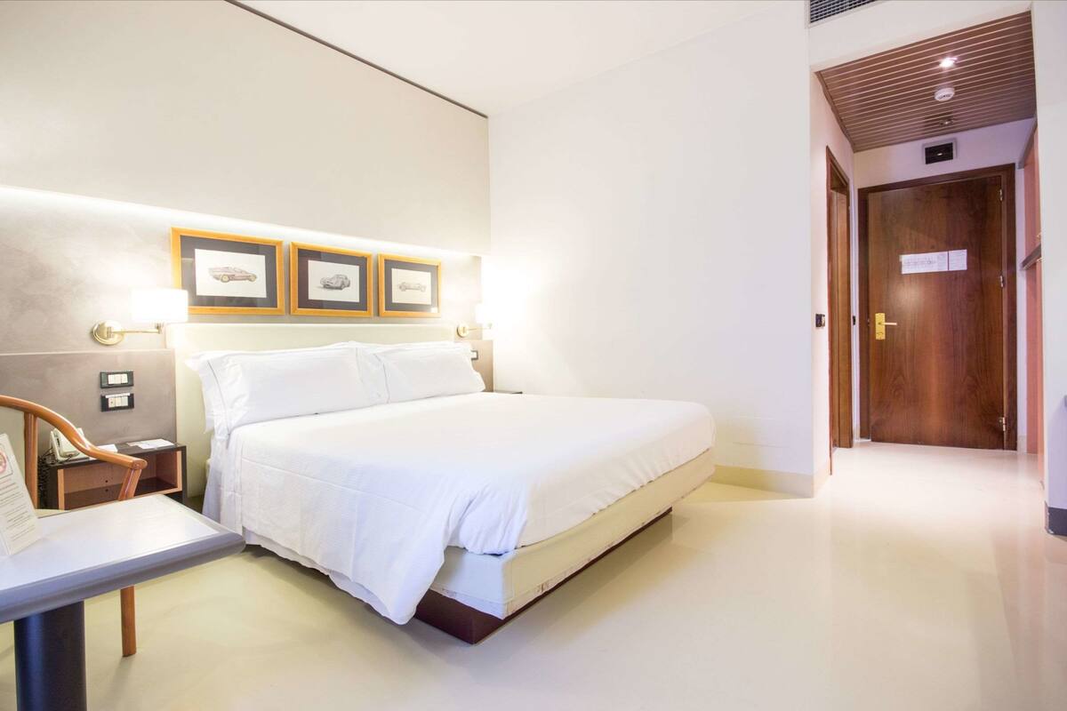 Ein Hotelzimmer mit Bett, Nachttisch, Lampe, gerahmten Bildern an der Wand und einer Holztür.