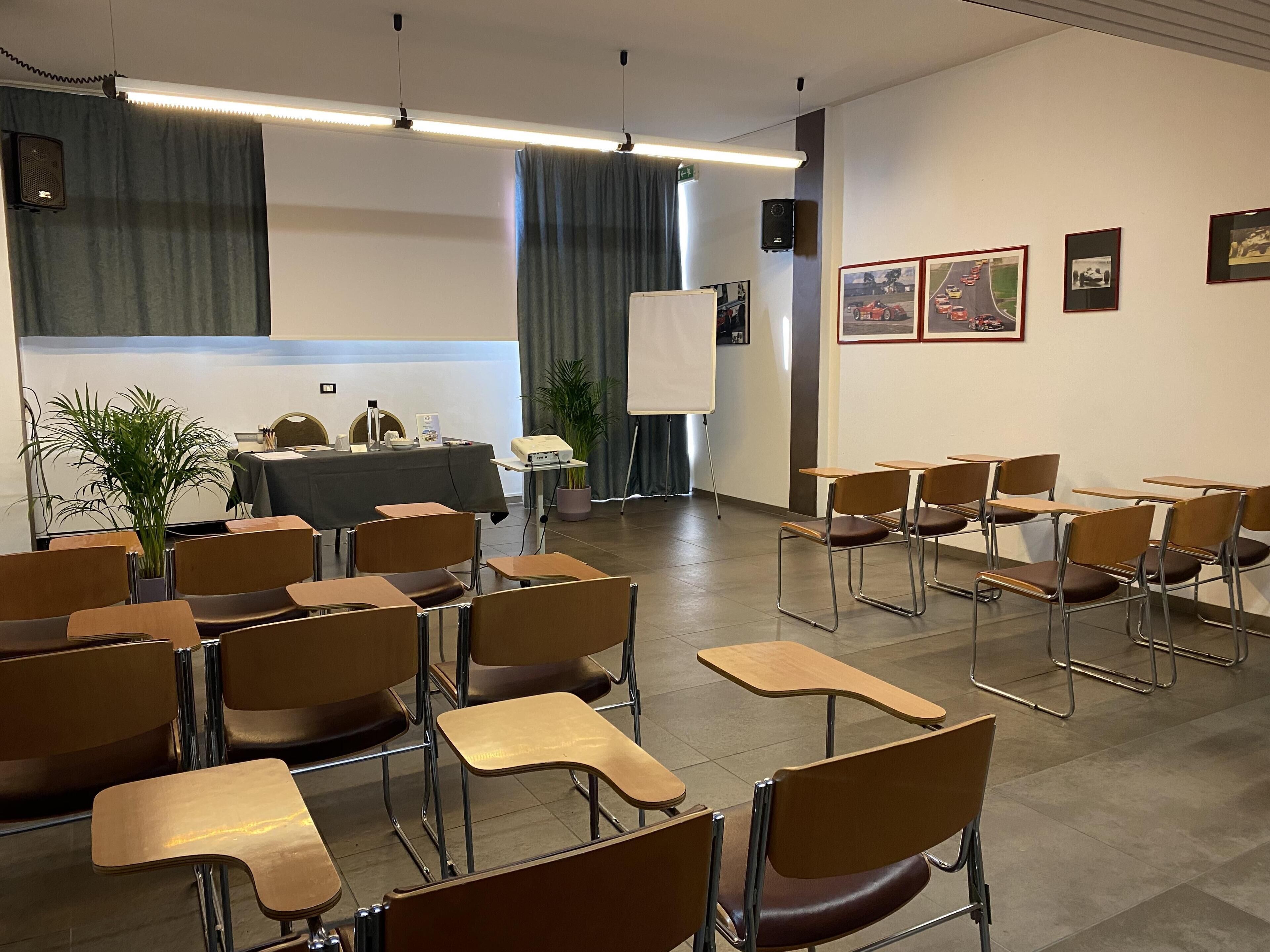 A classroom with desks and chairs arranged in rows, a projector, a whiteboard, and framed pictures on the wall.