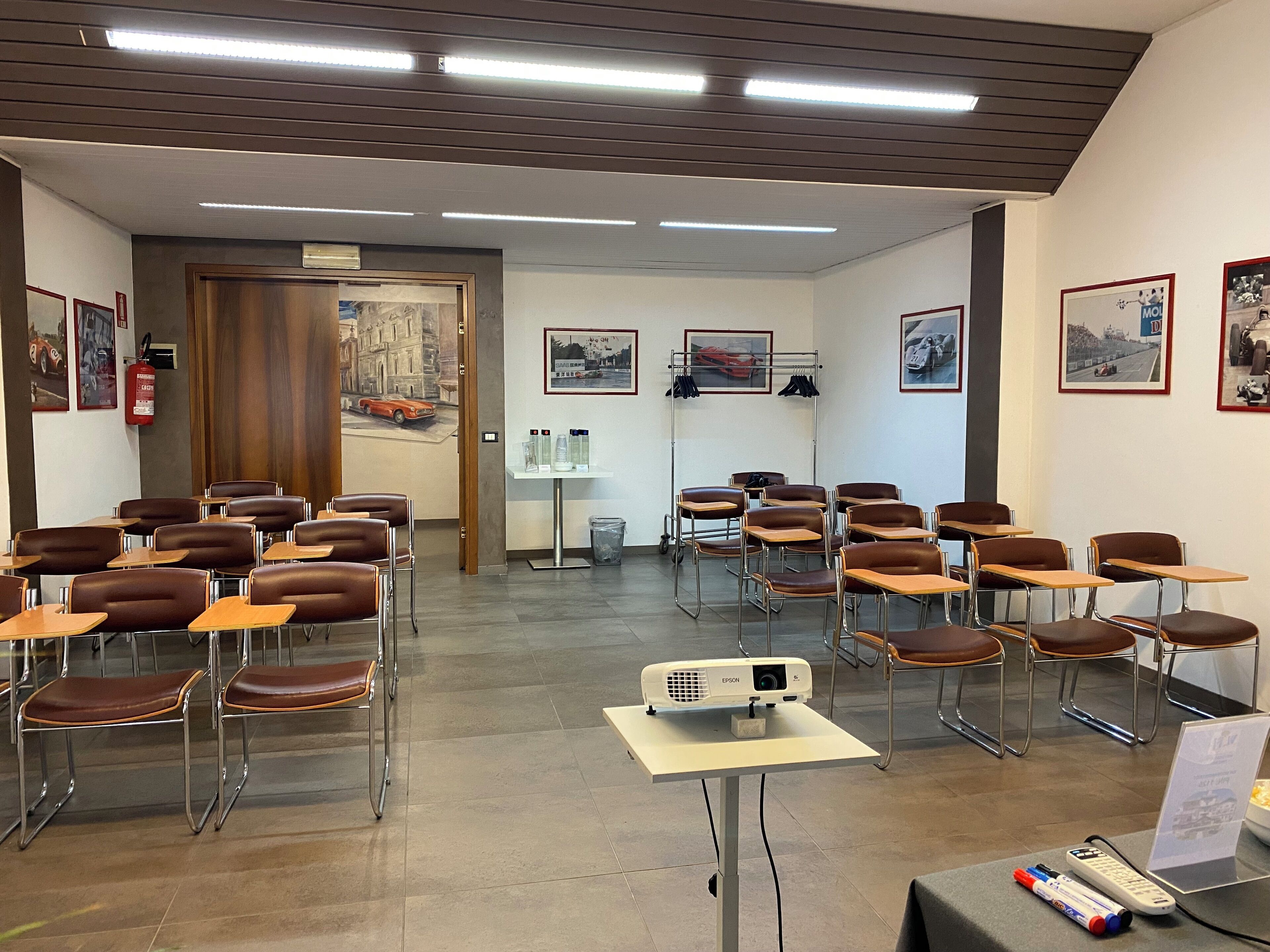 Un'aula con banchi e sedie disposti in file, un proiettore su un tavolo e quadri appesi alle pareti.