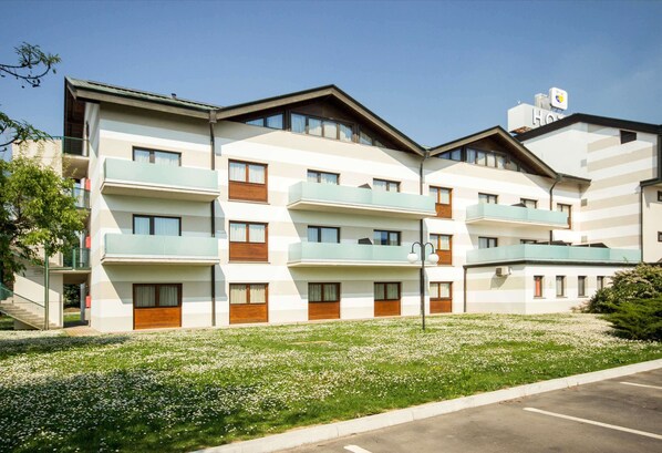 Un hotel de varios pisos con balcones, estacionamiento y una zona verde con flores blancas.