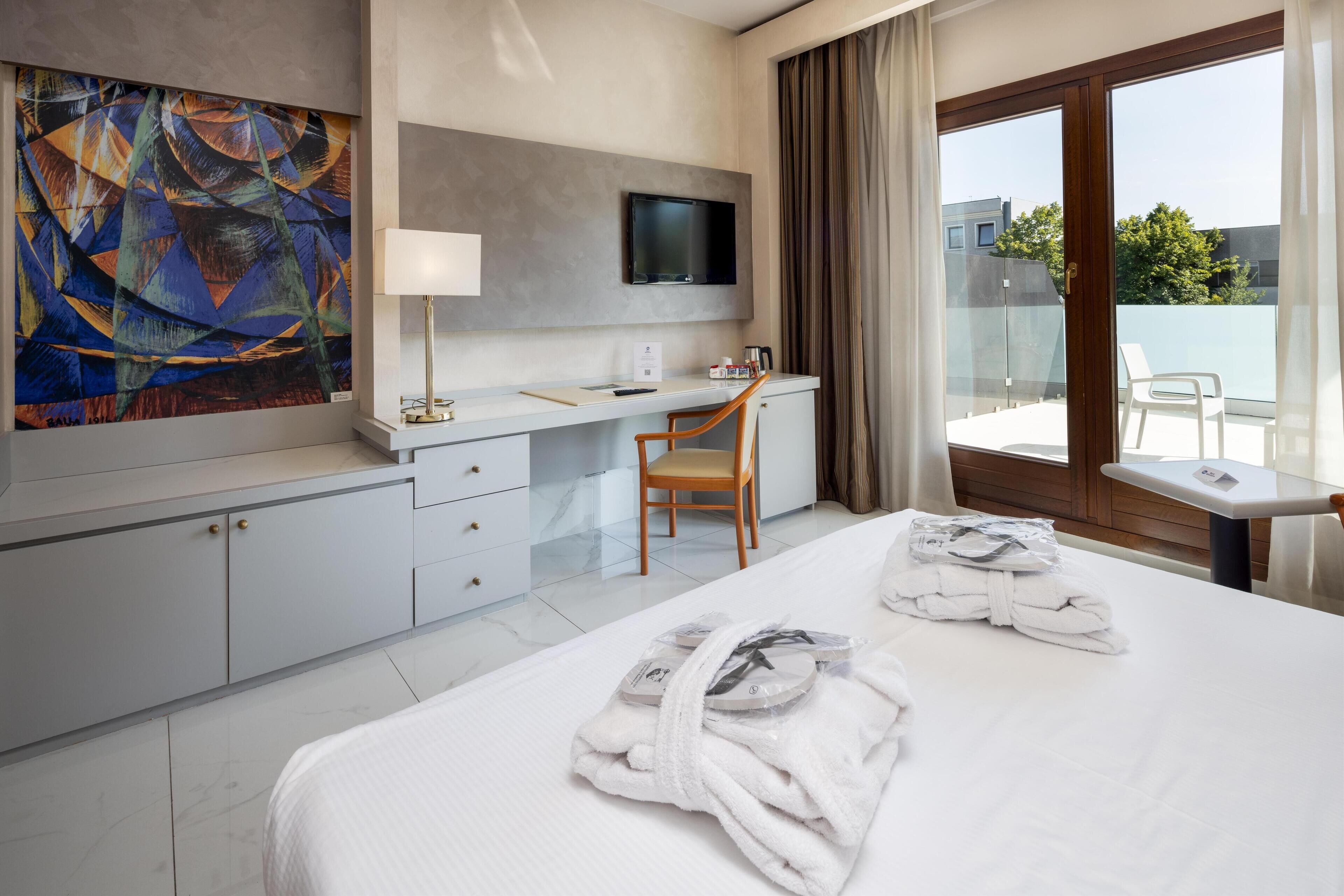 Ein Hotelzimmer mit Bett, Schreibtisch, Stuhl, Fernseher und einem großen abstrakten Gemälde an der Wand.