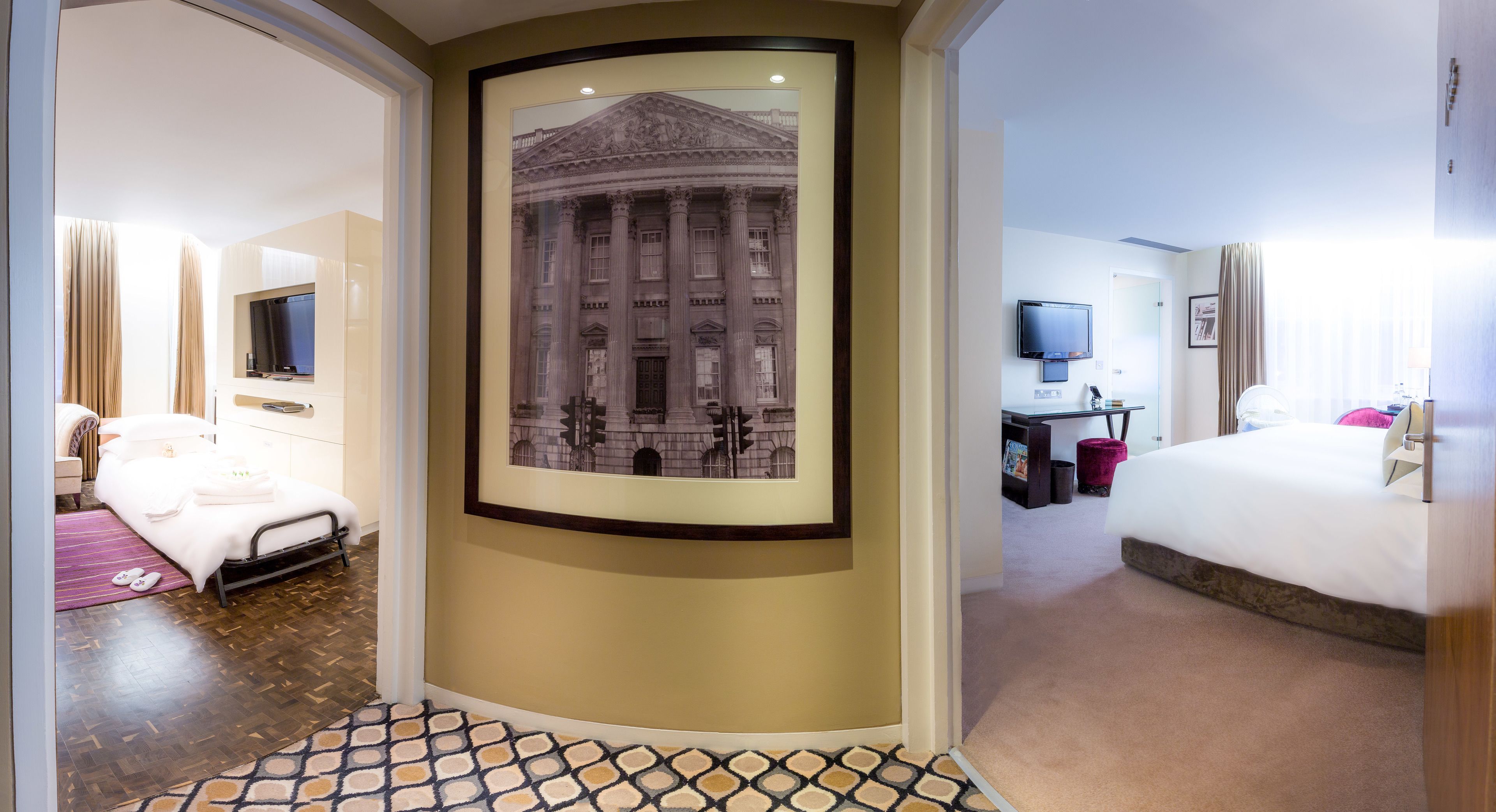 A hotel room with a large framed picture of a classical building, a bed, a desk with a computer, and a television.
