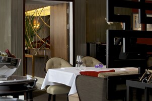 A dining area with a table set for two, a piano, and a plant in the background.